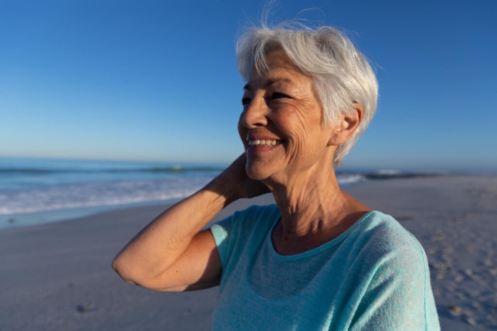 Mulher com pele madura sorrindo na praia em um dia ensolarado, representando saúde, bem-estar e cuidados com a pele no verão.