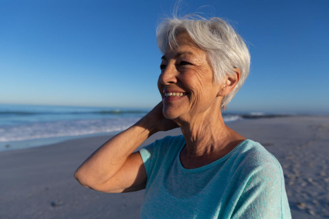 Mulher com pele madura sorrindo na praia em um dia ensolarado, representando saúde, bem-estar e cuidados com a pele no verão.